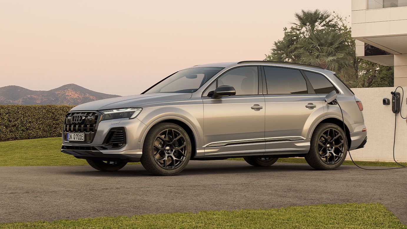 Bočný pohľad na strieborné Audi Q7 SUV TFSI e pri nabíjaní pri modernej vile. Okolo vozidla prechádza muž so ženou.
