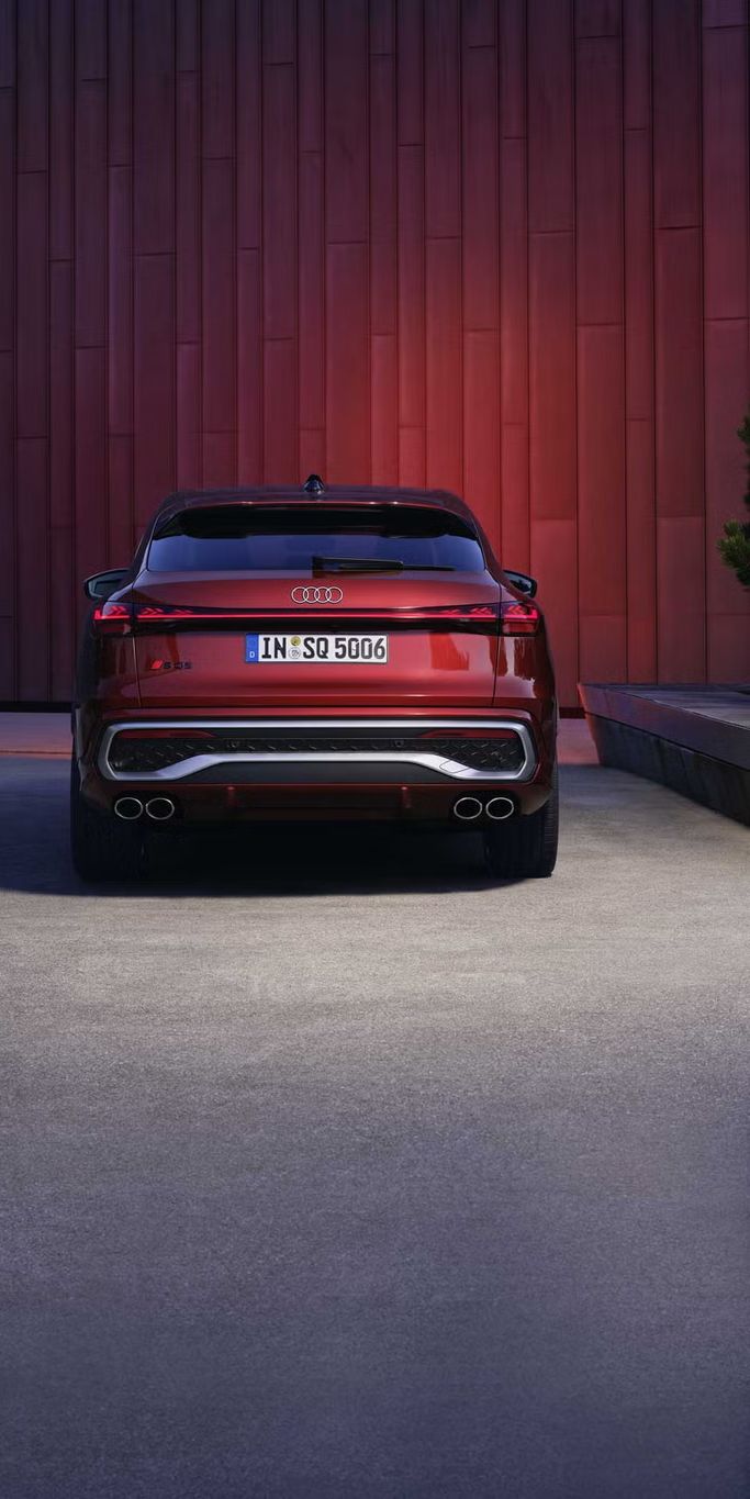 Heckprofil des roten SQ5 Sportback, welcher in einem Stadtpark vor einer Gebäudefassade steht und diese rot anleuchtet.