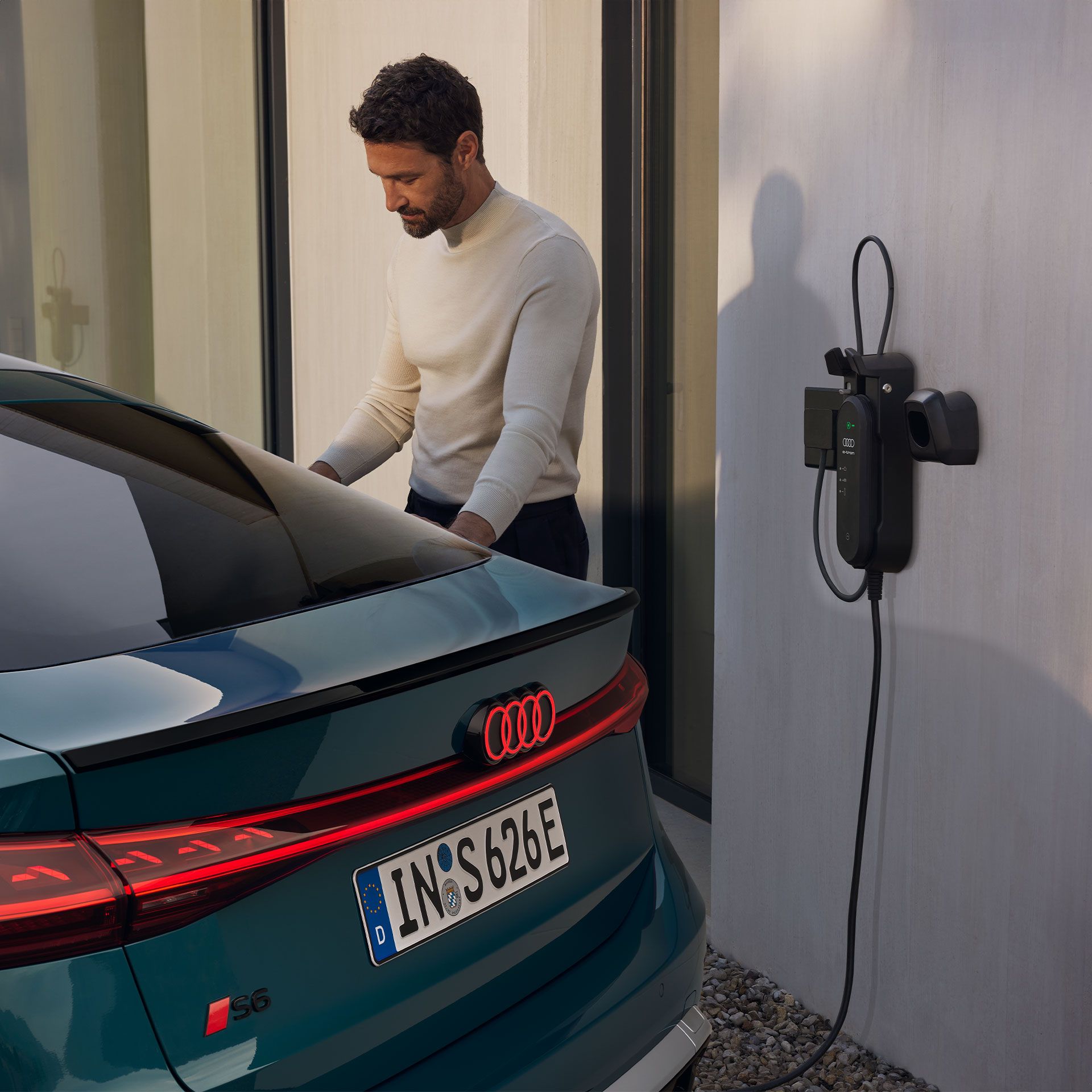 Pohľad na zadné partie Audi S6 e-tron zaparkovaný pri domácej nabíjacej stanici. Vedľa vozidla stojí muž a pripája ho k nabíjacej stanici.