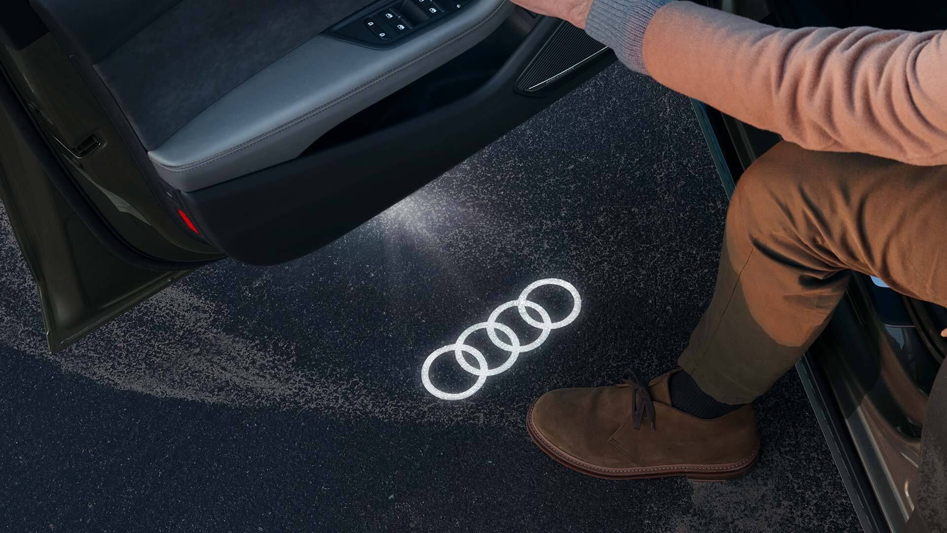 Muž sedí na strane vodiča v aute, drží otvorené dvere a pod dverami je na zemi logo Audi. Muž má obuté hnedé topánky.