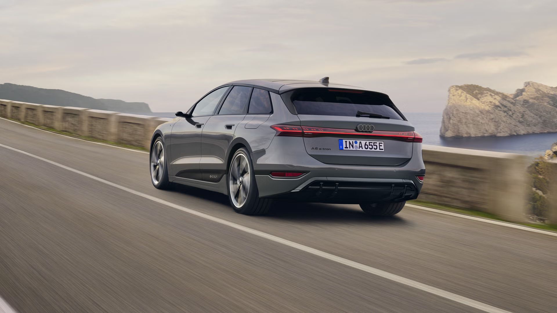 Šikmý pohľad na zadnú časť vozidla A6 Avant e-tron, ktoré jazdí po ceste pozdĺž mora. V pozadí sú viditeľné skaly a hory. 