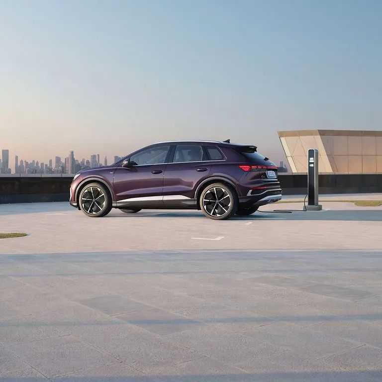 Audi Q4 e-tron zaparkovaný na strešnom parkovisku pri verenej nabíjacej stanici. Vozidlo je pripojené a nabíja sa. V pozadí je výhľad na centrum mesta.