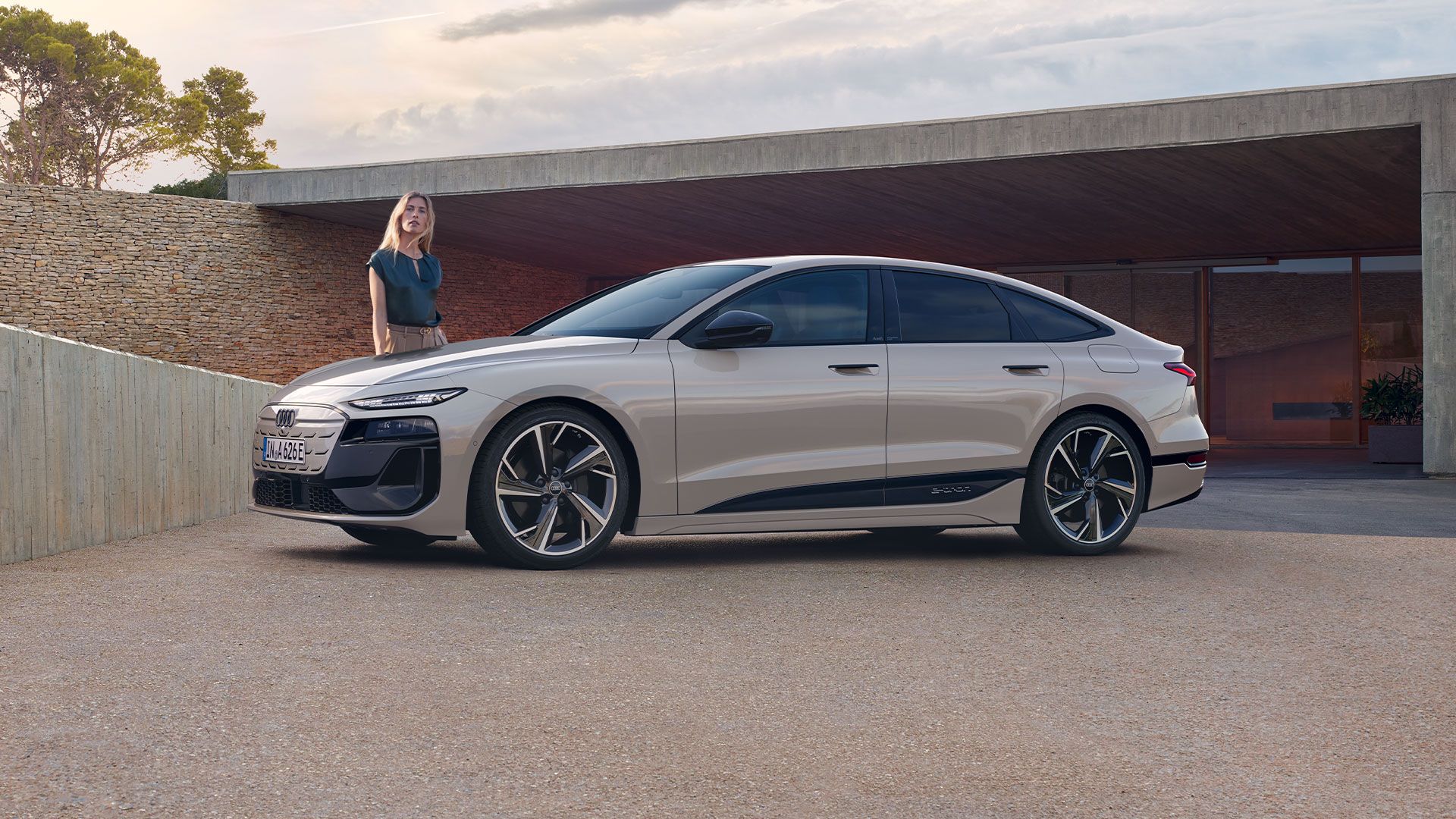 Elektrické Audi A6 Sportback e-tron zaparkované pred moderným domom pri západe slnka. Vľavo pri vozidle stojí mladá žena.