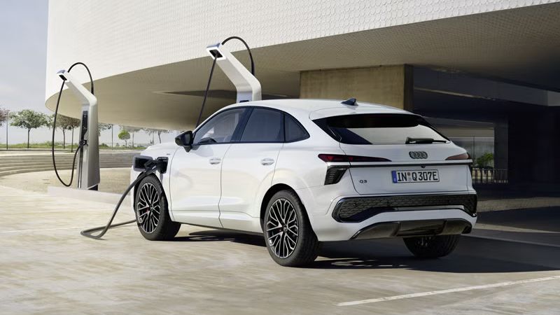 Biele Audi Q3 Sportback e-hybrid sa nabíja na nabíjacej stanici pred modernou budovou, vozidlo vrhá dlhé tiene.		