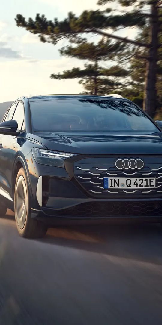 Audi Q4 e-tron počas dynamickej jazdy po ceste popri jazere z jednej strany a lese z druhej strany. V pozadí je výhľad na zalesnené kopce a jazero.