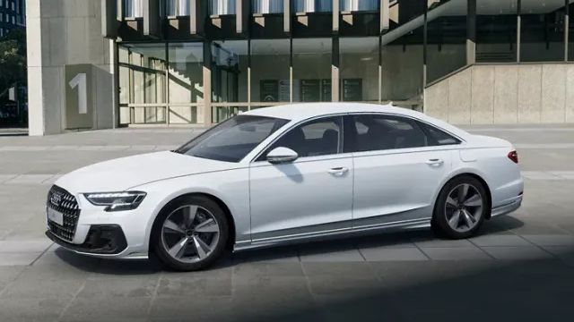 Biele, luxusné Audi A8 L TFSI e parkuje pred modernou budovou. Budova má vysoké okná a je z betónu.