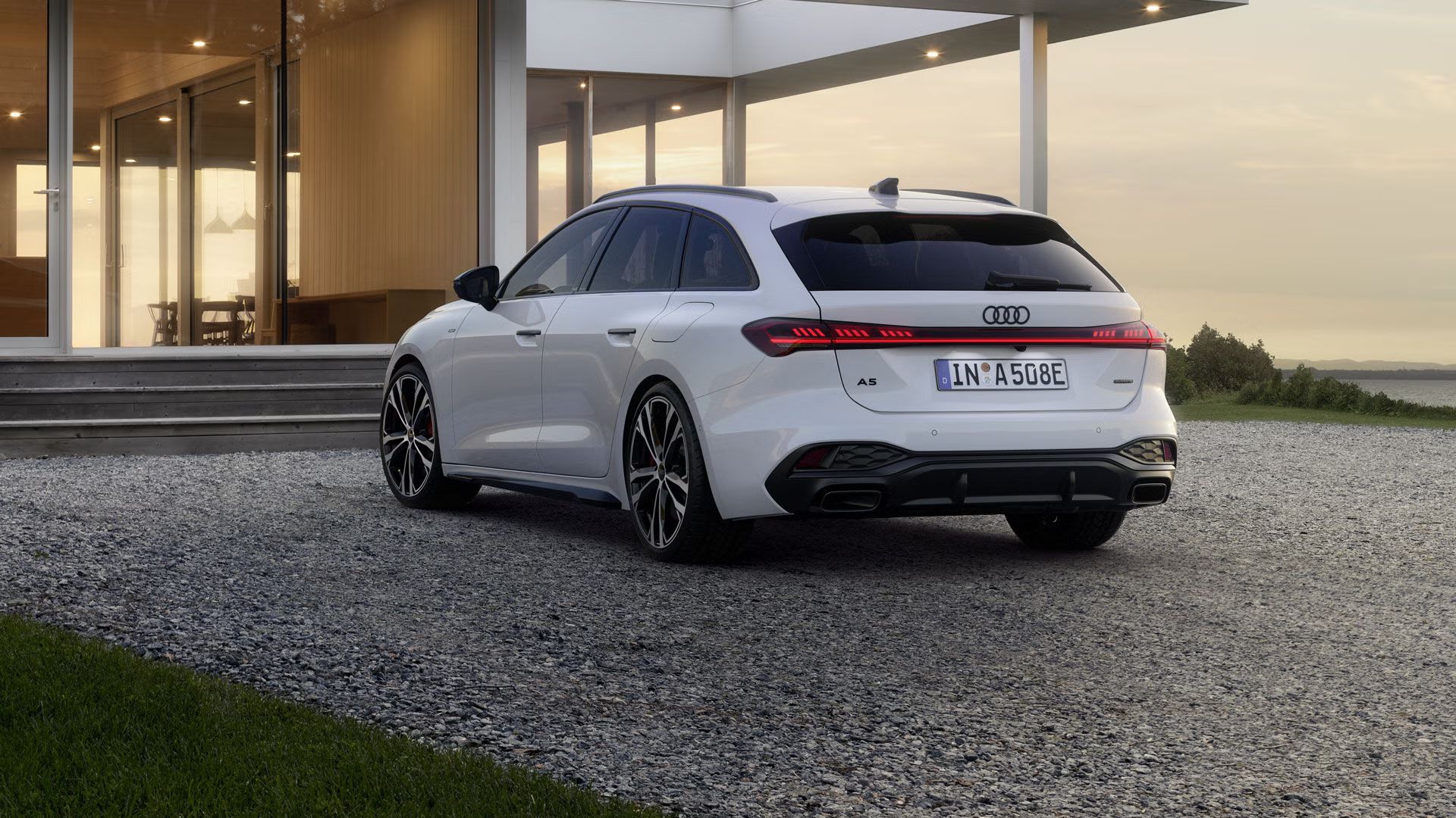 Der A5 Avant e-hybrid steht in einer schrägen Heckperspektive auf einer Kiesfläche vor einem modernen Wohnhaus.