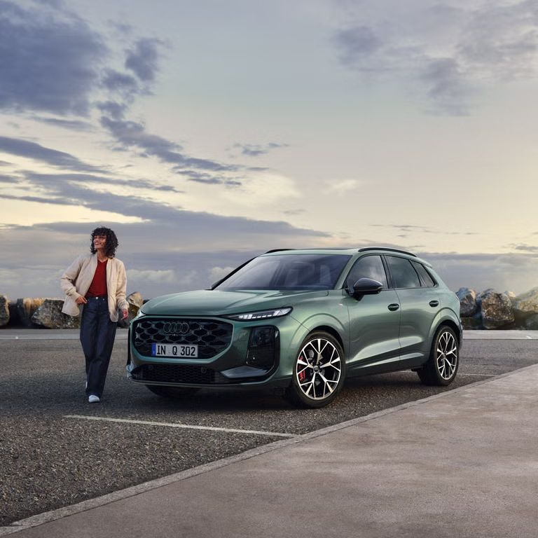 Seitliche, vordere Ansicht auf den geparkten Q3 SUV an einer Küstenstraße, daneben eine junge Frau. 