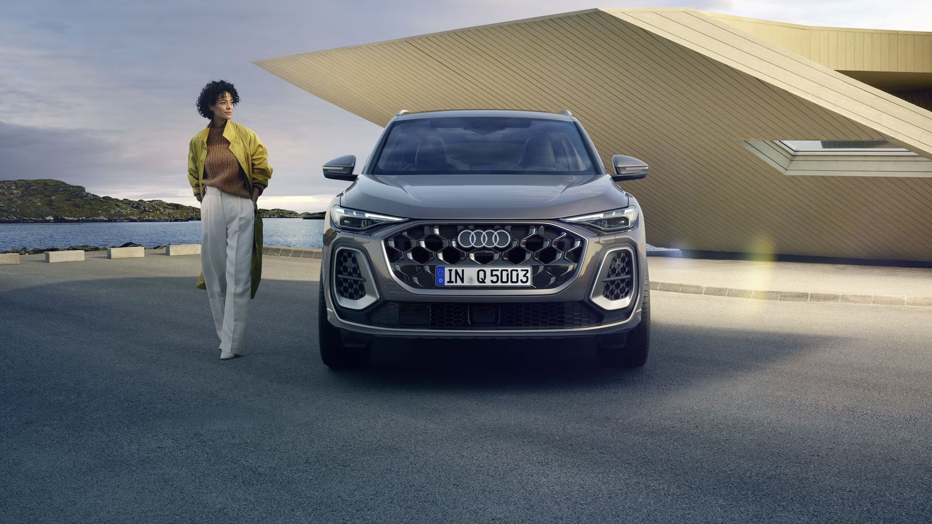 Audi Q5 SUV z čelného profilu na parkovisku priamo na pobreží, za ním malý futuristický dom. Vedľa Audi Q5 SUV stojí mladá, módne oblečená žena.
