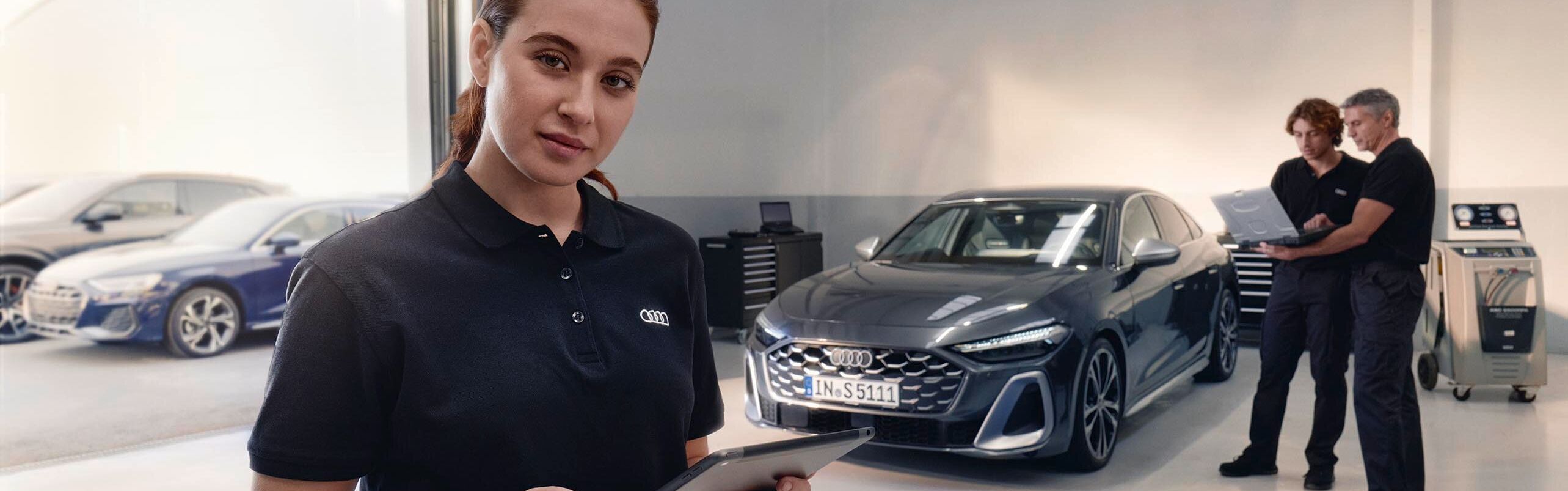 Servisná pracovníčka stojí v servise automobilov a drží tablet. V pozadí sú dvaja muži, ktorí sa rozprávajú pred zaparkovaným autom. Miestnosť je moderná a dobre osvetlená.