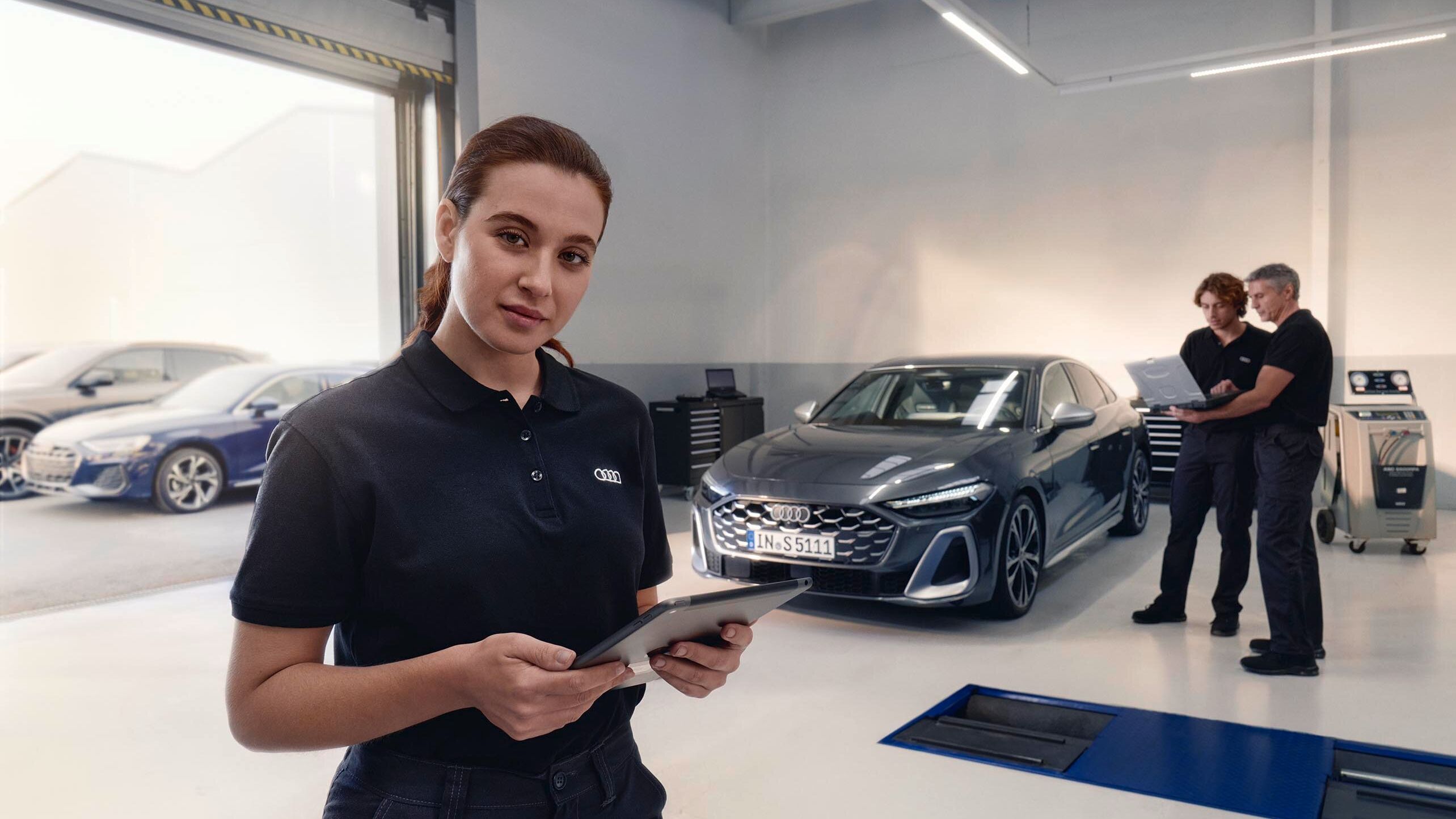 Servisná pracovníčka stojí v servise automobilov a drží tablet. V pozadí sú dvaja muži, ktorí sa rozprávajú pred zaparkovaným autom. Miestnosť je moderná a dobre osvetlená.