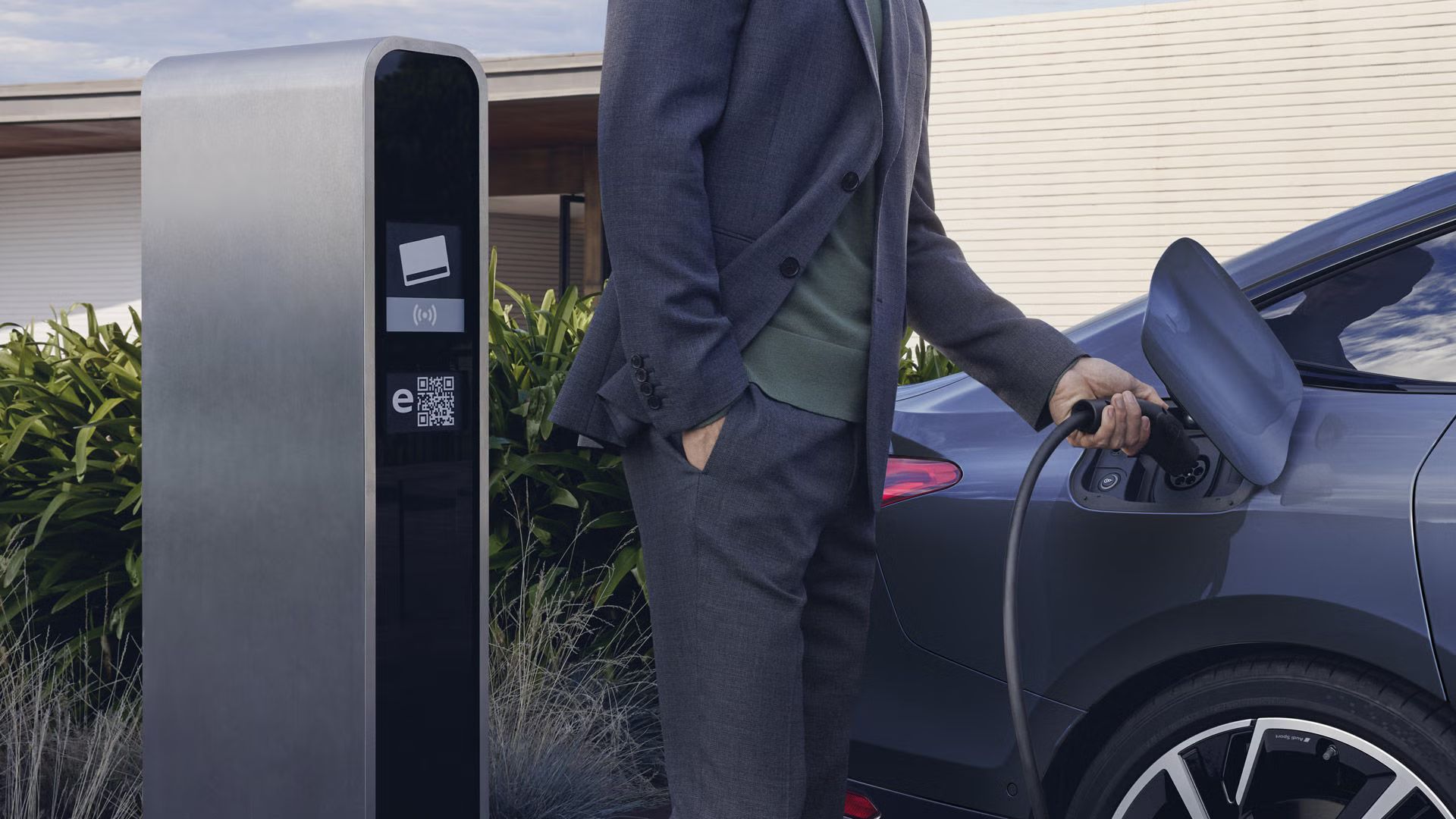 Detailný záber na nabíjací konektor modelu A6 Sportback e-tron, ku ktorému muž pripája nabíjací konektor. V pozadí je vidieť bielu modernú budovu. 