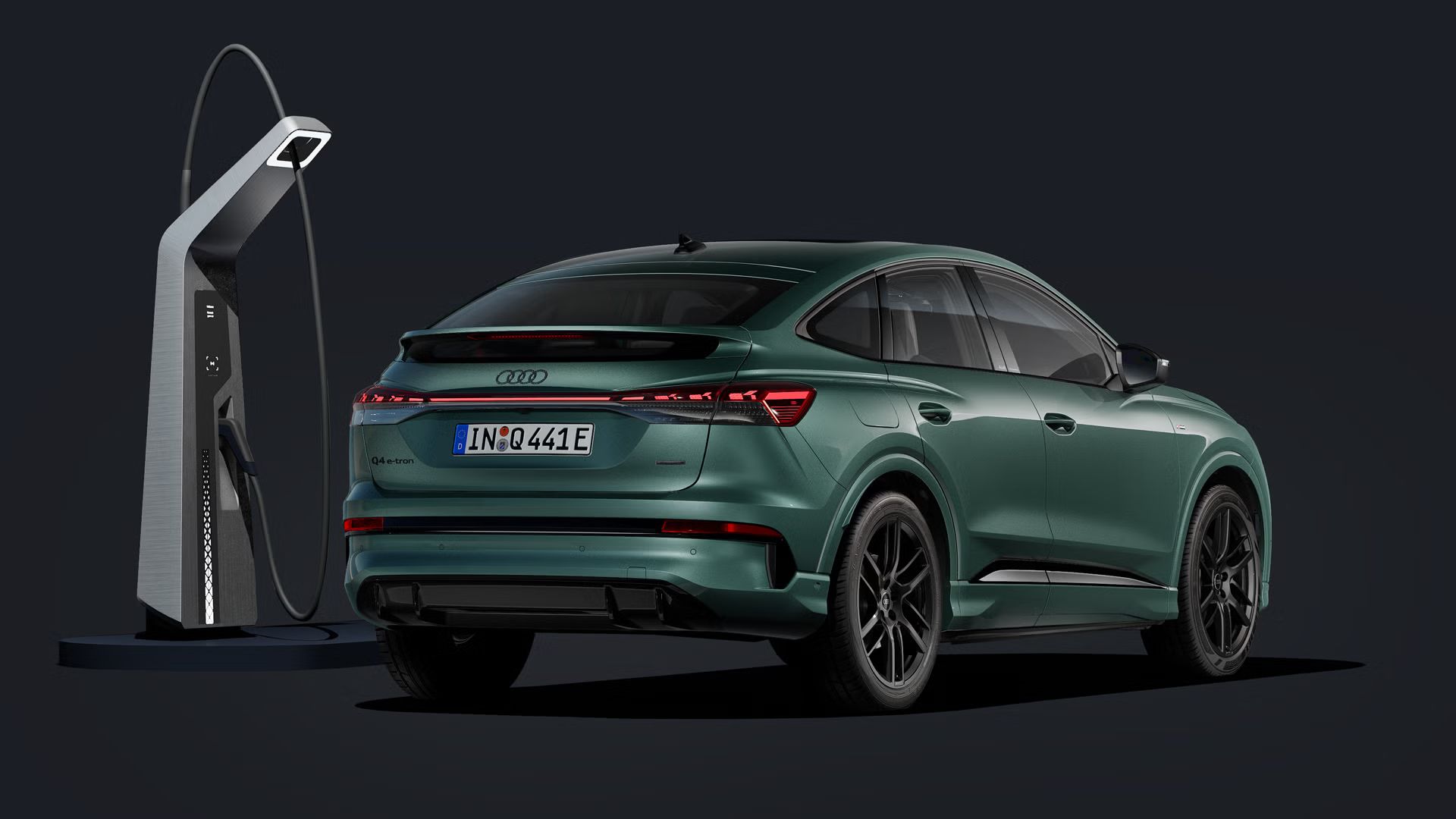 Pohľad na zadné partie Q4 Sportback e-tron šikmo z boku na tmavom pozadí, s modernou nabíjacou stanicou vedľa vozidla.