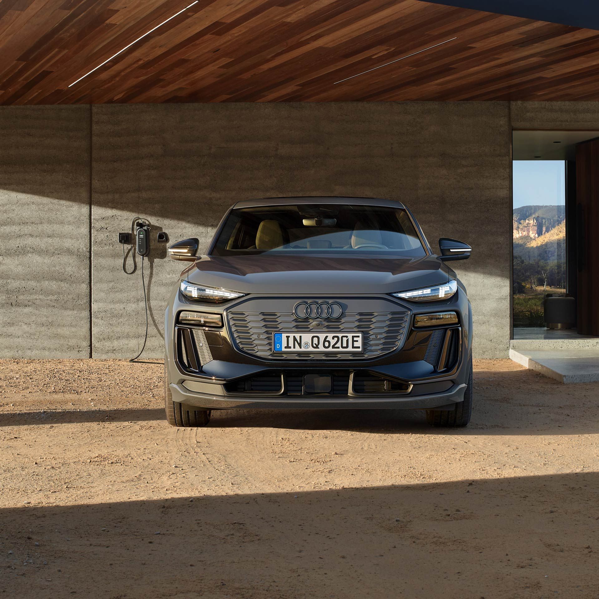 Pohľad spredu na Audi Q6 Sportback e-tron zaparkované pred moderným domom. Vľavo vedľa neho je na stene nainštalovaná domáca nabíjacie stanica.