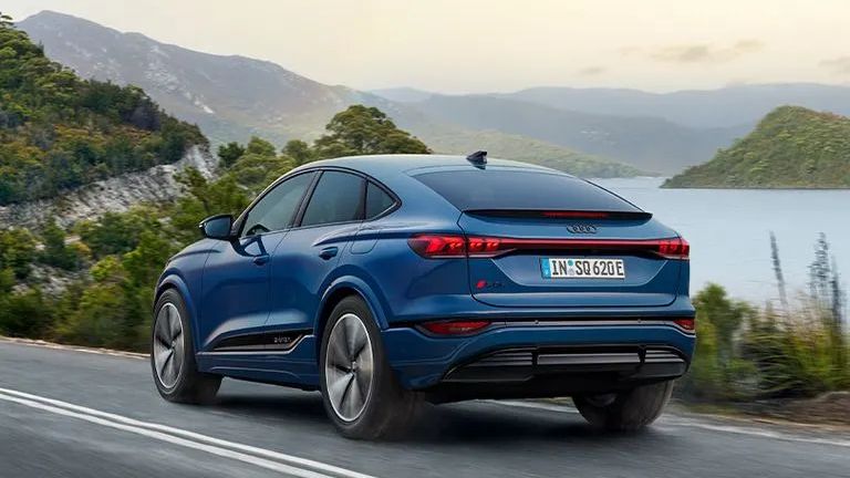 Modré Audi Q5 prechádza po ceste pri jazere. Cestu lemuje zeleň, vedľa cesty je veľké jazero. V pozadí sú hory. Obloha je jasná, s jemnými mrakmi.