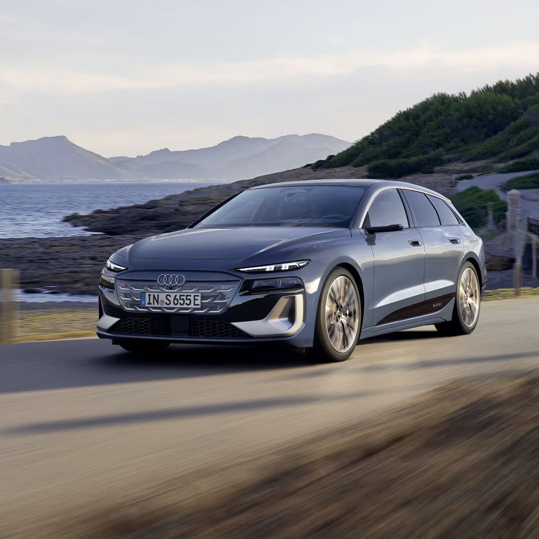 Audi S6 Avant e-tron z čelného pohľadu počas dynamickej jazdy po ceste pozdĺž pobrežia. V pozadí sú vidieť hory a vodnú hladinu. 