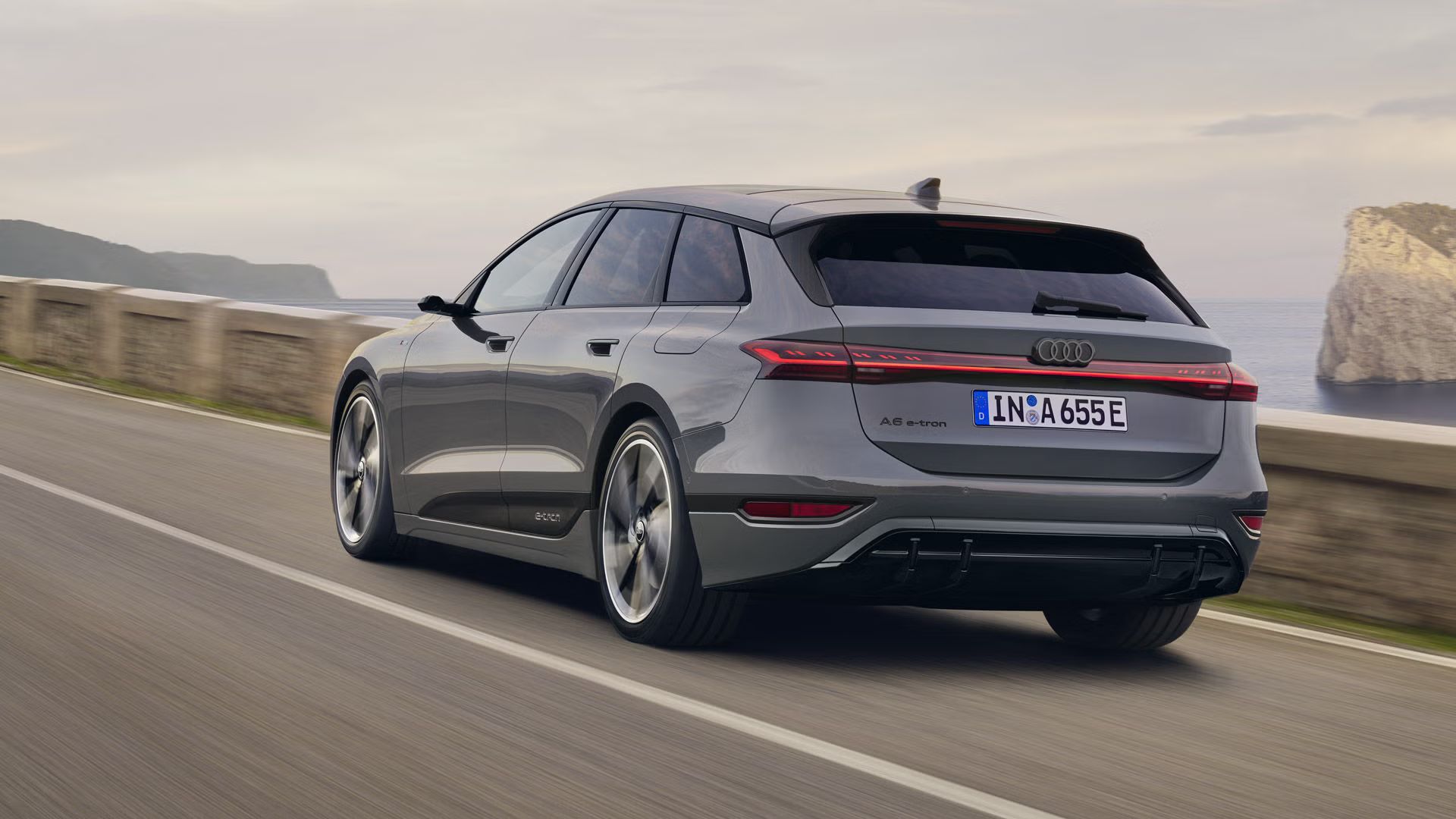 Šikmý pohľad na zadnú časť vozidla A6 Avant e-tron, ktoré jazdí po ceste pozdĺž mora. V pozadí sú viditeľné skaly a hory. 