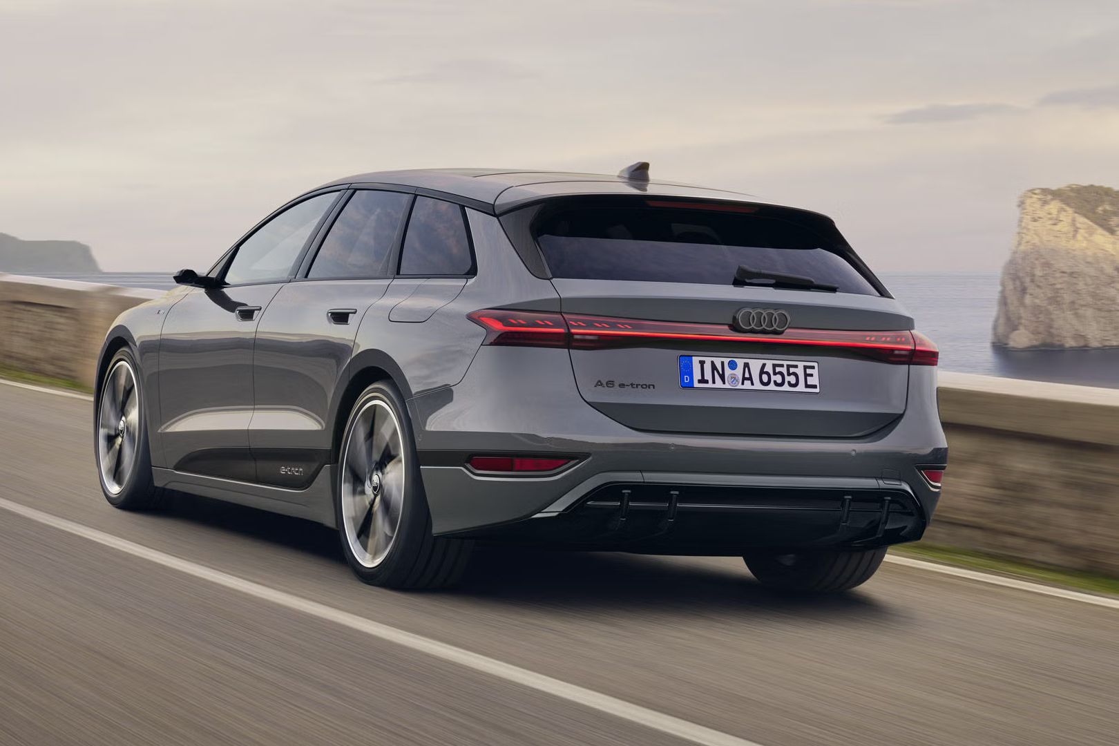 Šikmý pohľad na zadnú časť vozidla A6 Avant e-tron, ktoré jazdí po ceste pozdĺž mora. V pozadí sú viditeľné skaly a hory. 