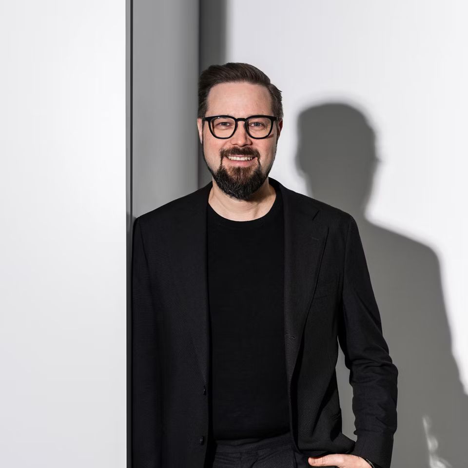 Usmievajúci sa muž v čiernom tričku a čiernom obleku sa opiera o stenu a pozerá sa priamo do objektívu.